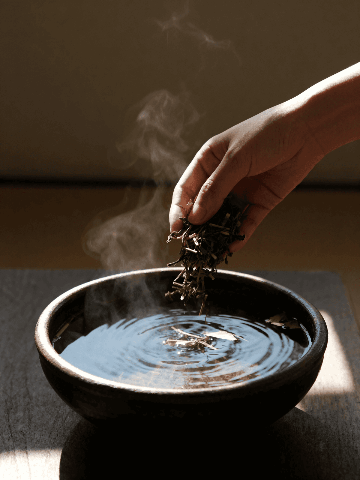 Hands placing herbs into a dark ceramic bowl of steaming water